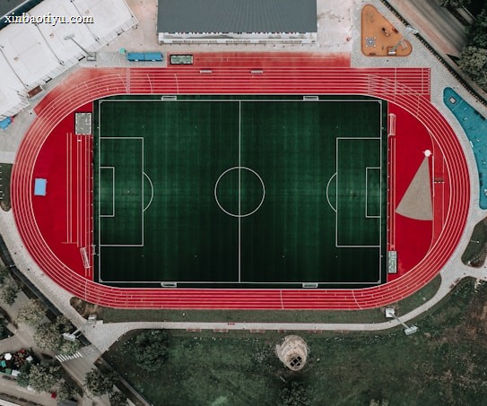 An aerial view of a large sports complex featuring a central green football field surrounded by a red running track. Adjacent to the track, there is a parking area, buildings, and pathways. Nearby, there are additional sports courts in different colors, including a blue swimming pool and orange basketball courts.