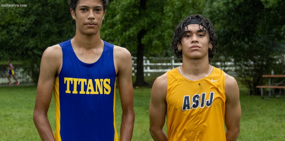 Two young athletes wearing sleeveless sports tops stand on a grassy field. The athlete on the left is wearing a blue top with the word 'TITANS' while the one on the right wears a yellow top with 'ASIJ'. The background features green trees and a white fence, with a bench visible on the right.
