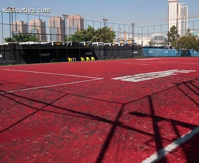 A sports court with a red playing surface and white lines, surrounded by a black mesh fence. In the background, tall buildings are visible, along with some trees and a few yellow chairs positioned near the fence.