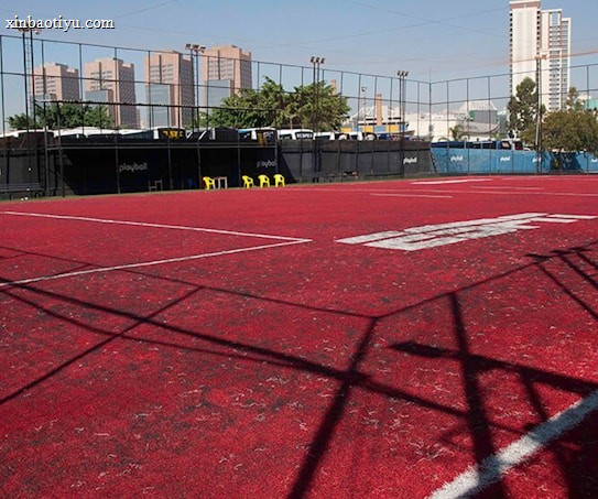 A sports court with a red playing surface and white lines, surrounded by a black mesh fence. In the background, tall buildings are visible, along with some trees and a few yellow chairs positioned near the fence.